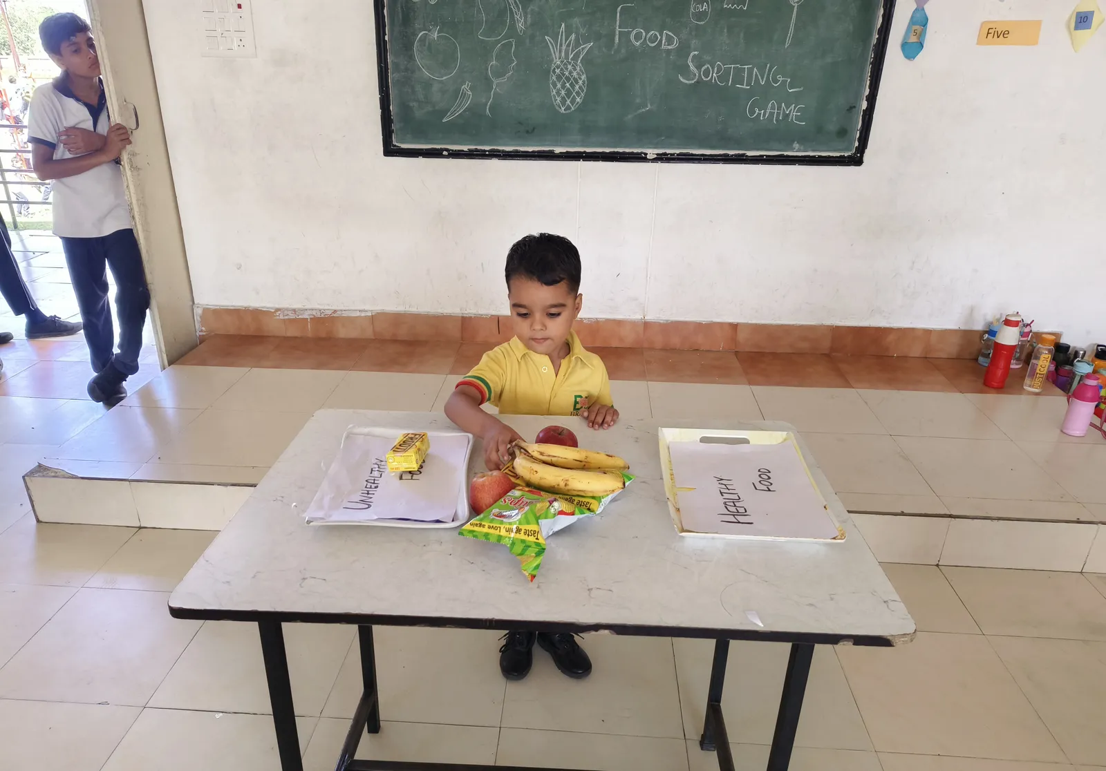 A boy sorts the food at the table, the game’s chalkboard on the wall behind him