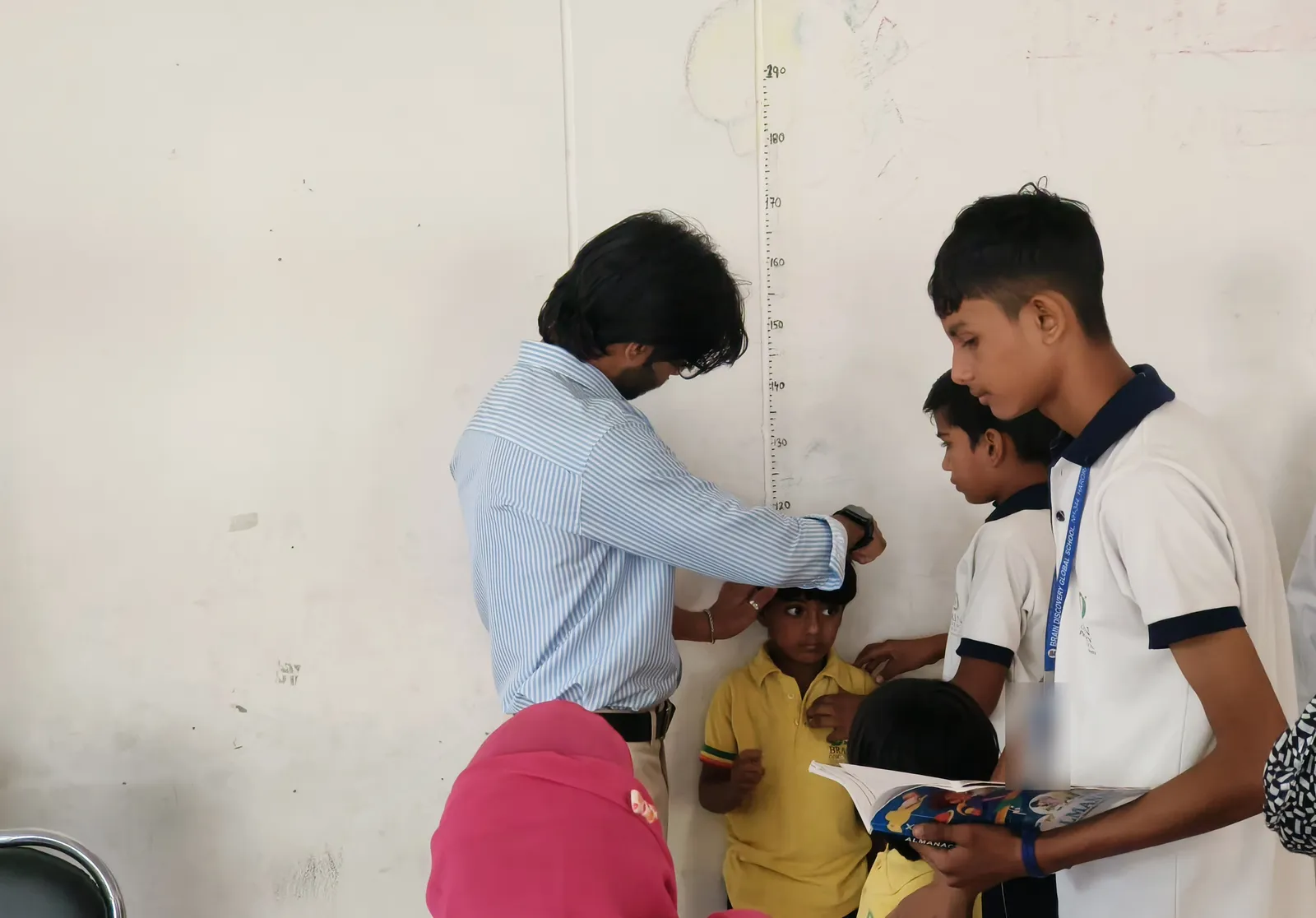 A teacher measures a boy’s height against the chart on the wall as older students help
