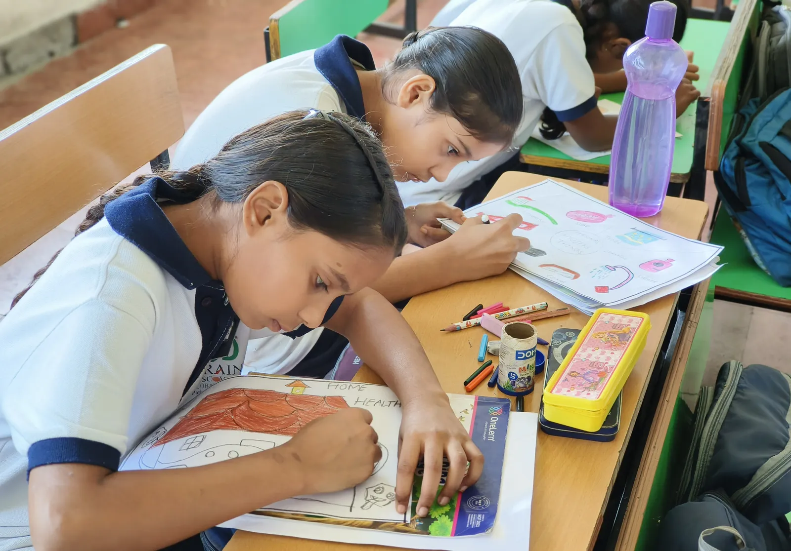 Two girls colouring their posters, one of a house titled Home Health, the other of a toothbrush, soap, towel and comb around the words Personal Hygiene Things