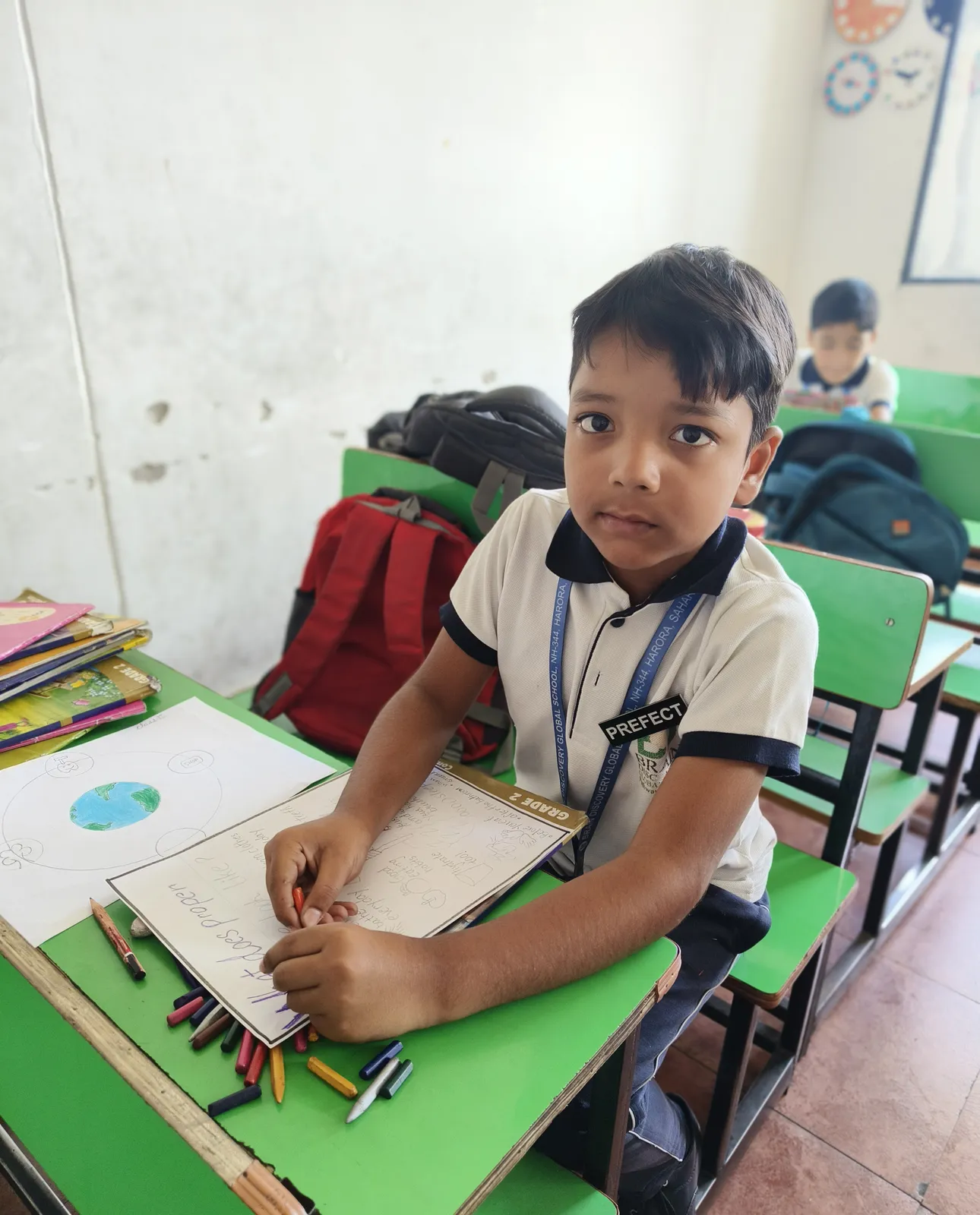 A Class 2 boy looks up from his worksheet, crayons spread across the desk beside a drawing of the Earth