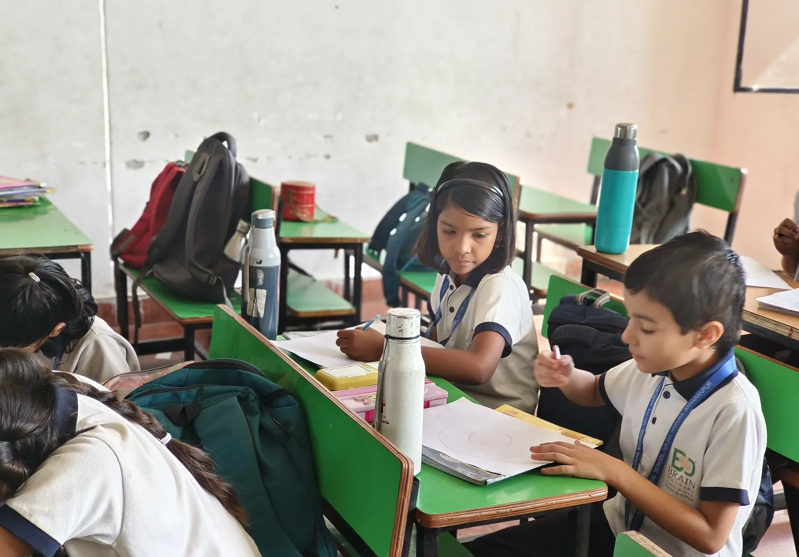 A girl and a boy drawing at their desks, water bottles and bags around them