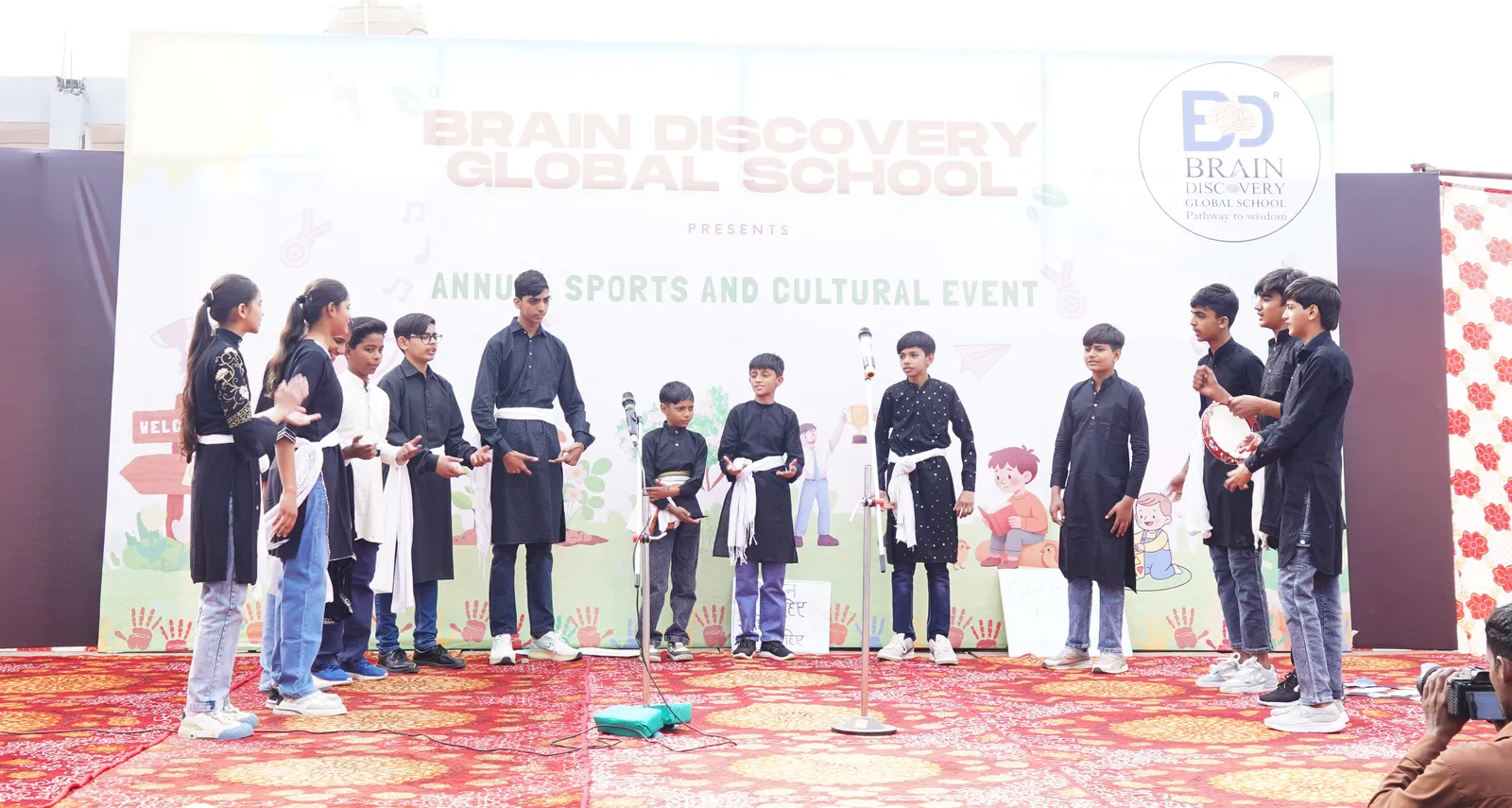 The cast in black kurtas and white sashes lined up across the stage, two boys with a tambourine on the right