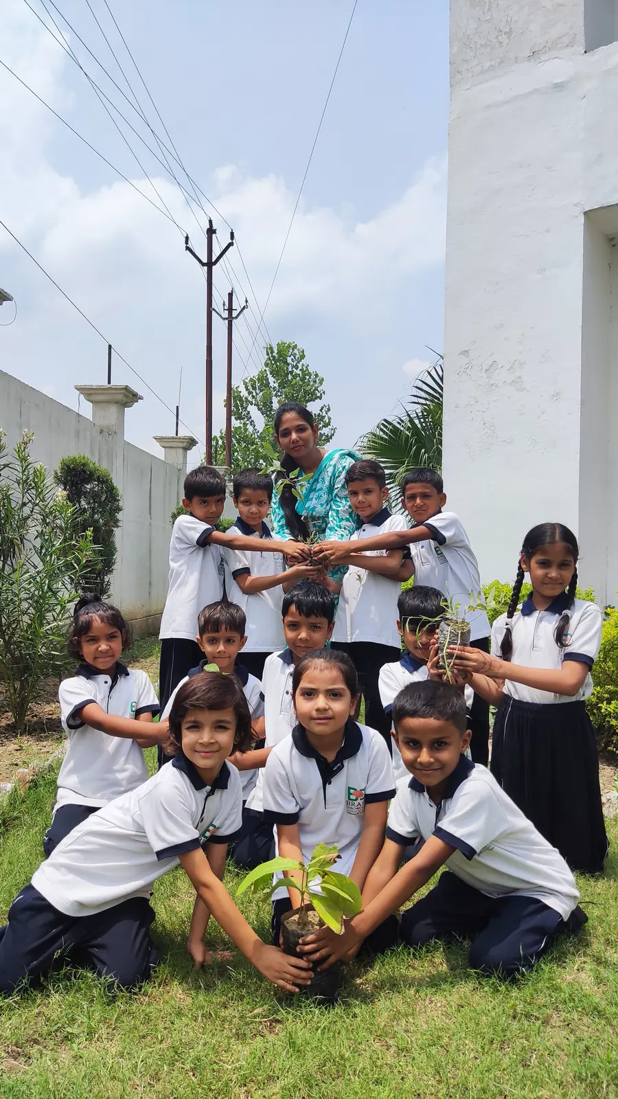 Young students reaching in together to hold one sapling, their teacher behind them